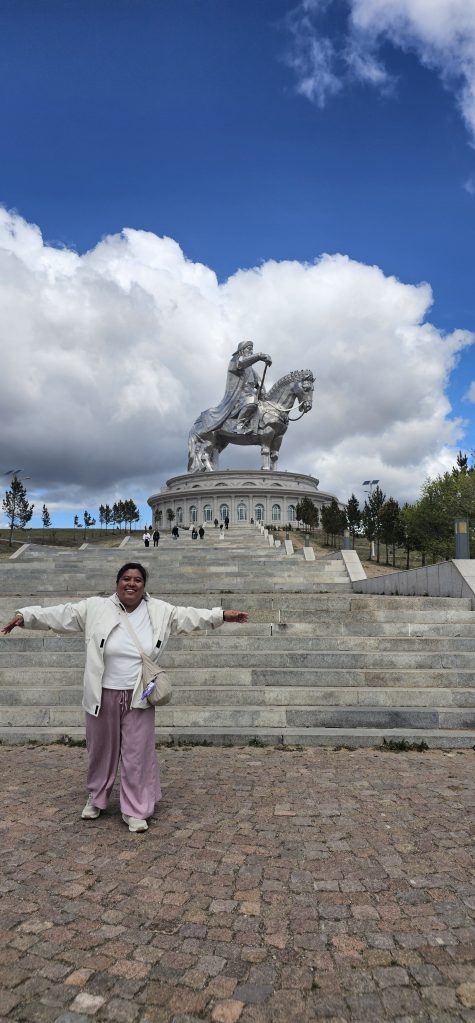 A smiling person stands with arms open in front of a large silver statue of a horse and rider on a pedestal against a backdrop of blue skies and clouds.