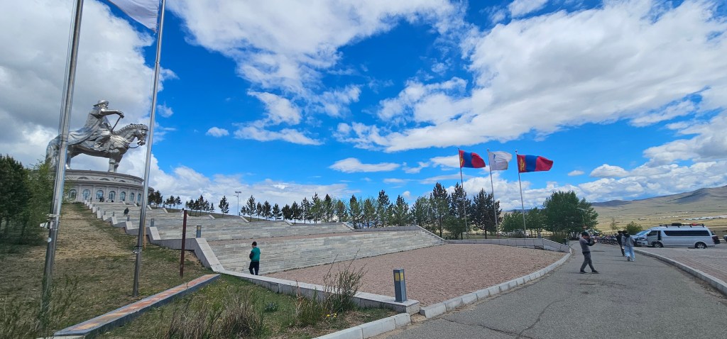 A panoramic view of a memorial site featuring a statue of a horseman, surrounded by trees and flags. The sky is partly cloudy with a vibrant blue tone.
