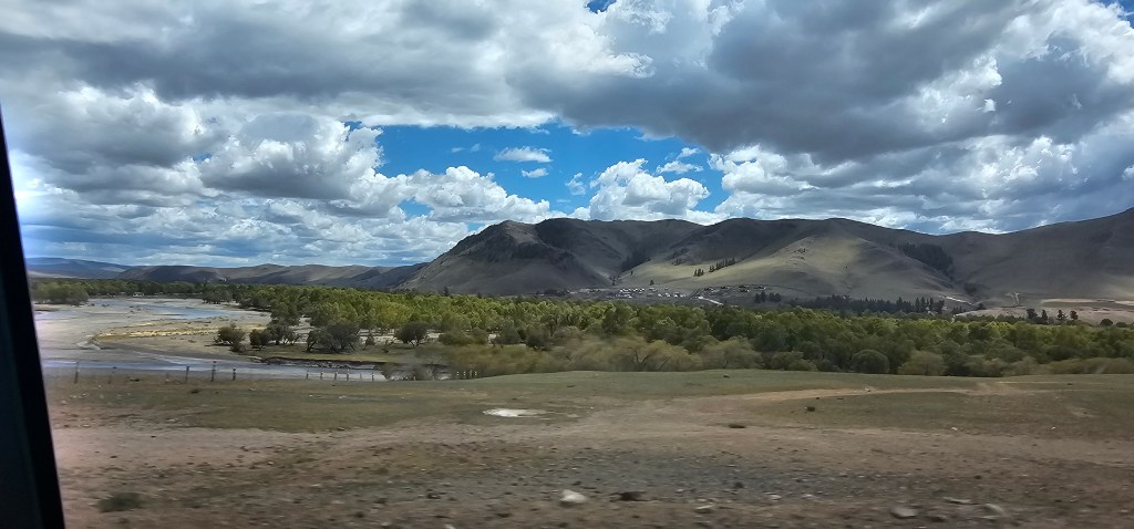 Scenic view of rolling hills with a river and forested area under a partly cloudy sky.