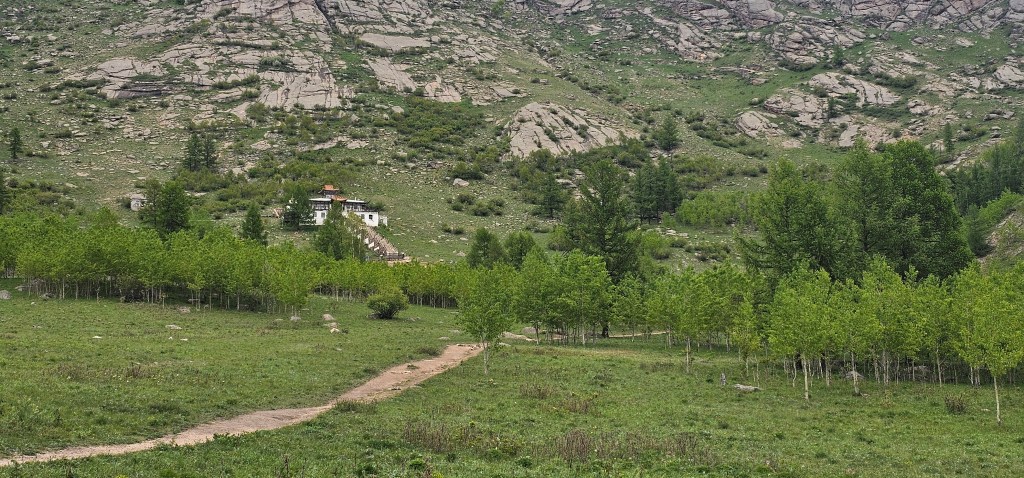 A scenic landscape featuring a winding dirt path leading through lush green fields and trees, with a small building nestled in the background against a rocky hillside.