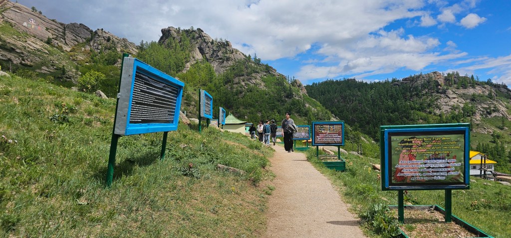 A scenic path with informational signs on either side, surrounded by green vegetation and hills under a partly cloudy sky.