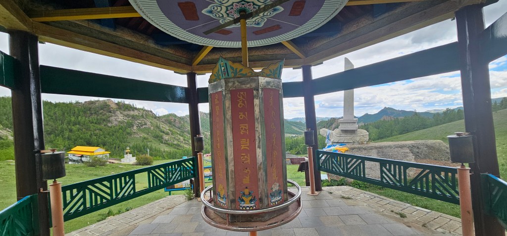 Buddhist prayer wheel in a pavilion, surrounded by mountains and green landscape.