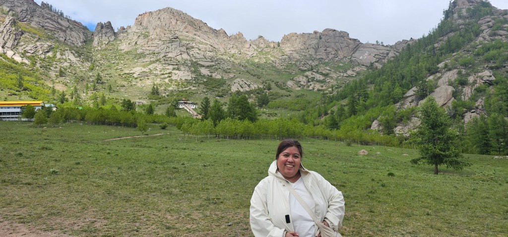 A smiling woman wearing a light-colored jacket stands in a grassy valley surrounded by mountains and trees.
