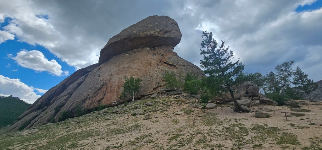 A large rock formation resembling a hat balanced on top of a massive boulder, surrounded by sparse vegetation and a cloudy sky.