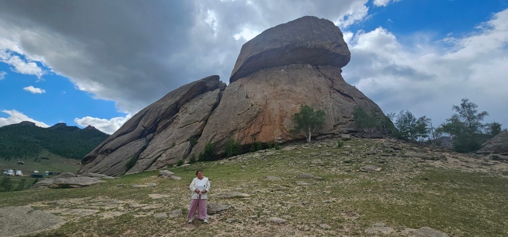 A person standing in front of a large rock formation under a cloudy sky, surrounded by grassy terrain and distant mountains.