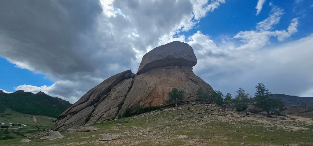 A large, flat-topped rock formation rises above a grassy landscape under a cloudy sky, with trees nearby and hills in the background.