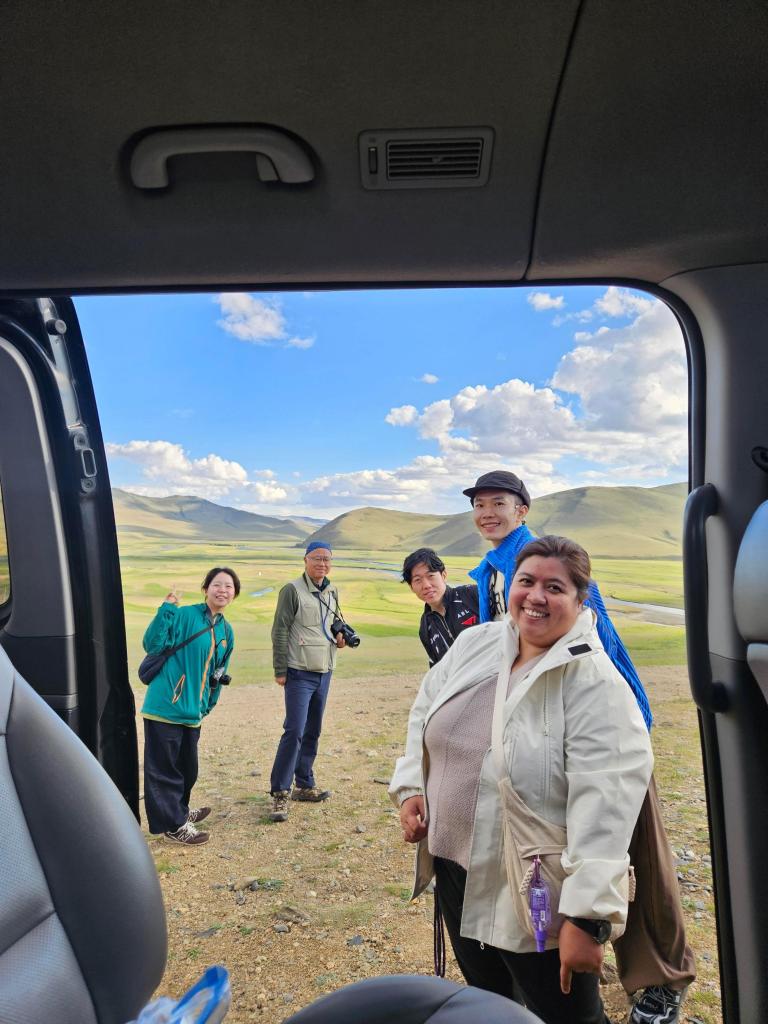 Group of five people smiling and posing at an outdoor location with rolling hills and a blue sky in the background, seen from inside a vehicle.