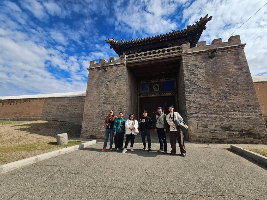 Group of five people standing in front of a large brick wall and a traditional ornate building entrance with a blue sky and clouds in the background.