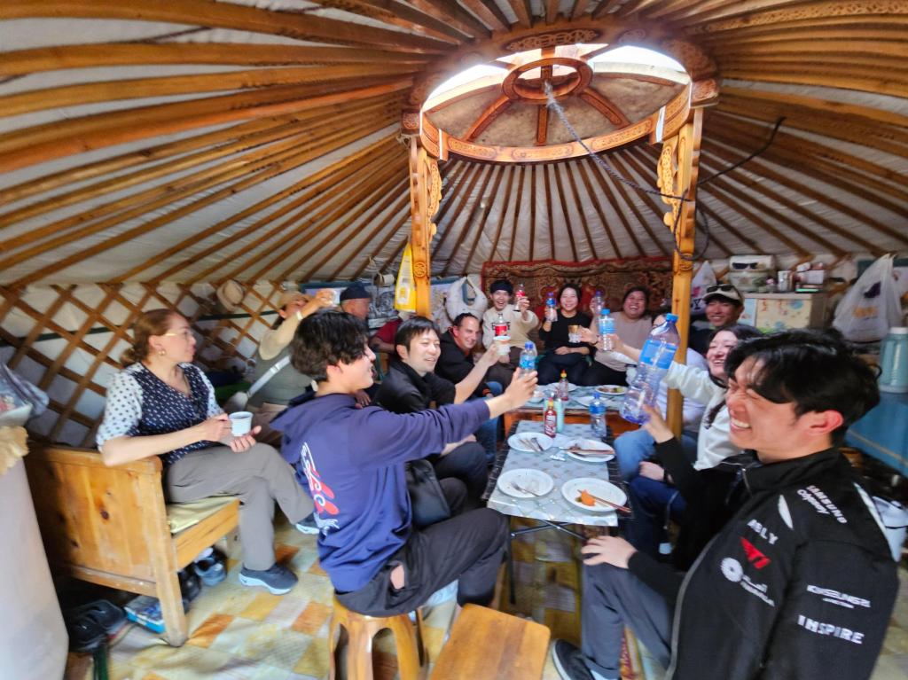 A group of people smiling and toasting with drinks inside a traditional yurt. The interior features wooden beams and a circular opening at the top, and there are plates of food on the table.