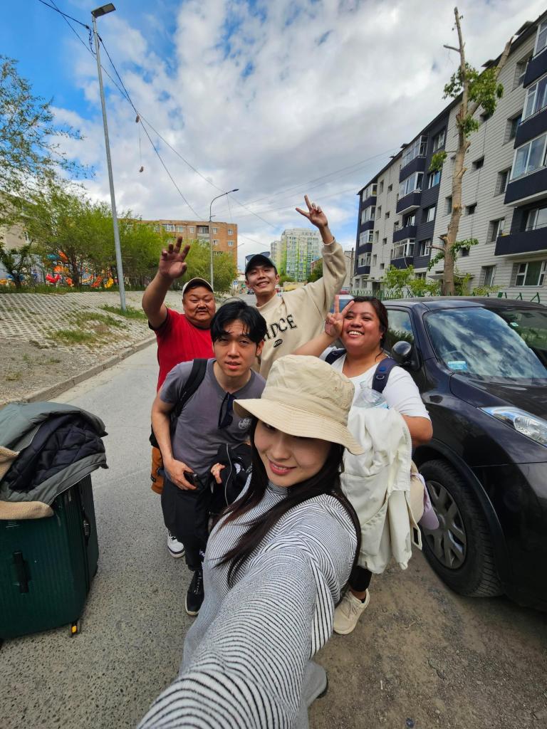 Group selfie of five people outdoors standing in a street, smiling and posing with peace signs. Luggage is visible in the foreground, and buildings are in the background under a cloudy sky.