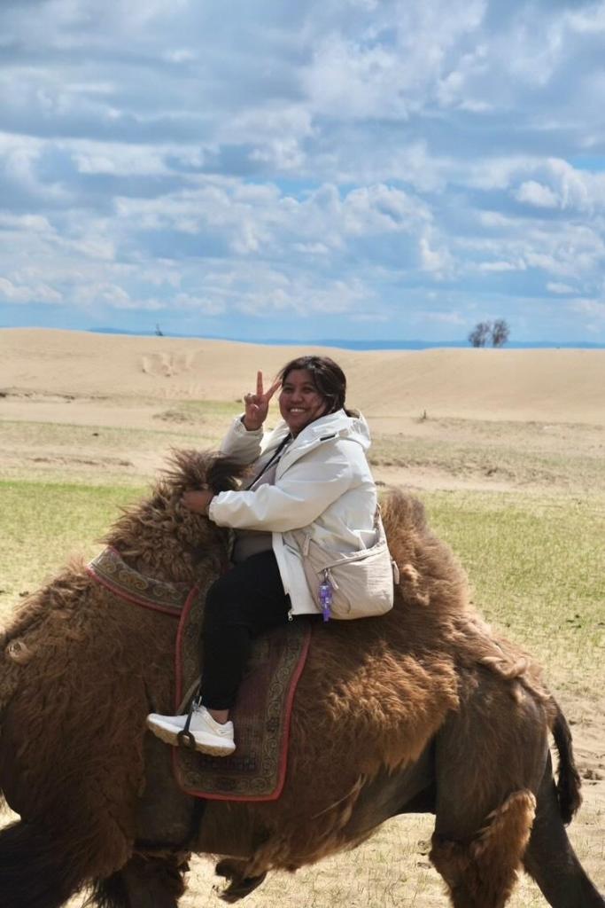 20260605_154026 A person riding a large, furry animal in a desert landscape, smiling and making a peace sign with one hand, under a partly cloudy sky.