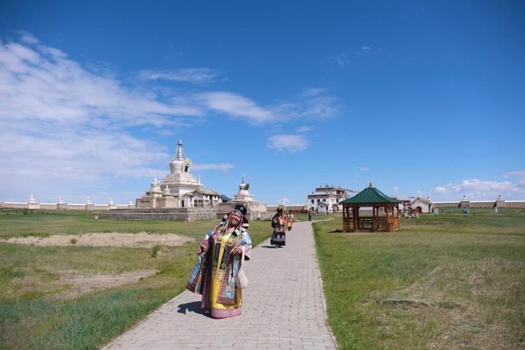 A group of people wearing traditional clothing walking along a stone pathway in a grassy landscape with a large, ornate building and a gazebo in the background under a blue sky.
