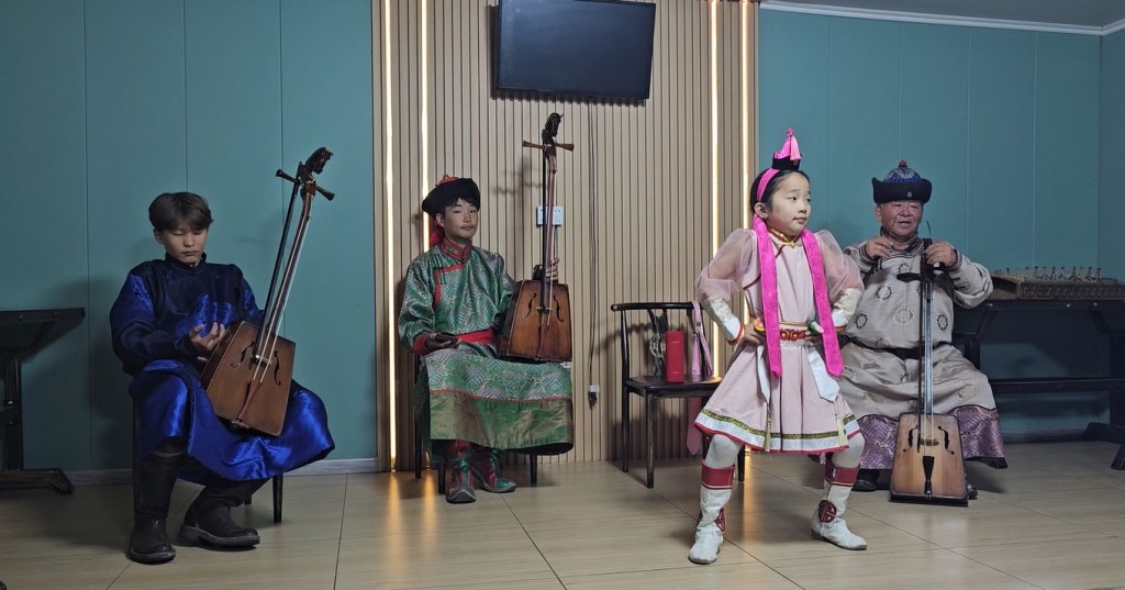 Four performers in traditional attire, playing Mongolian instruments and dancing in a cultural setting.